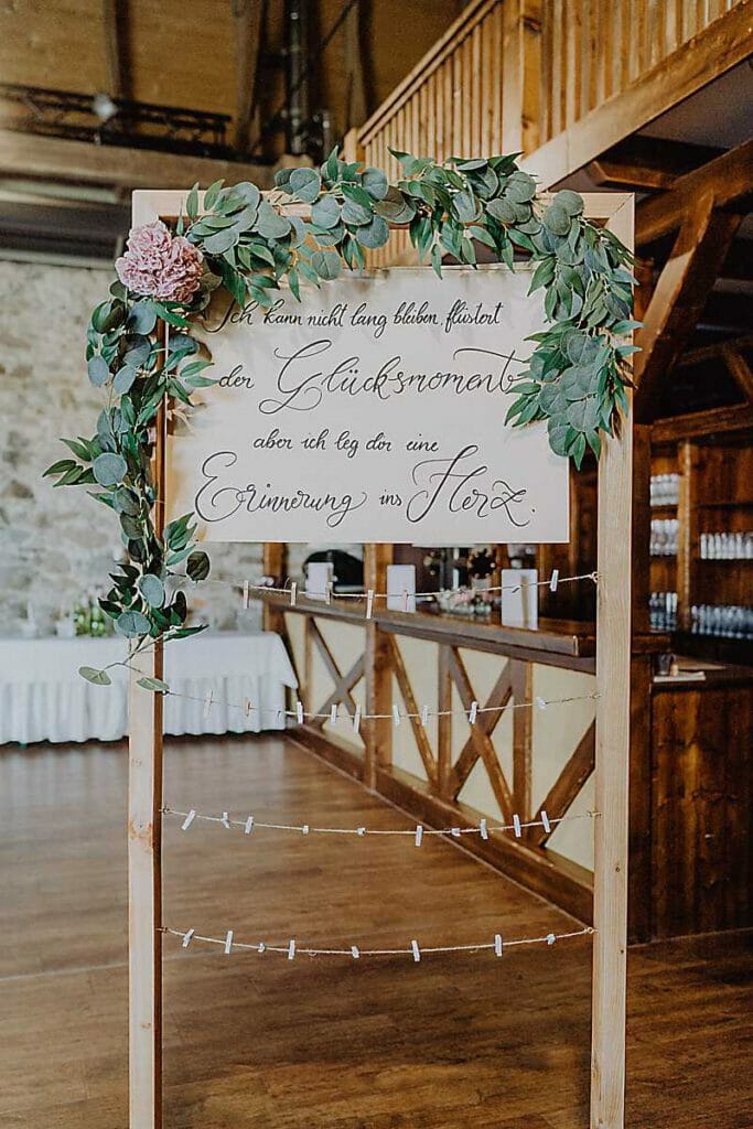 Ein mit grünen Blättern und einer rosa Blume verziertes Holzschild zeigt die elegante deutsche Kalligrafie einer Hochzeitsplanerin in Leipzig und Usedom. Unten hängen Schnüre mit Wäscheklammern in einer rustikalen Halle mit Holzbalken und Steinwänden.