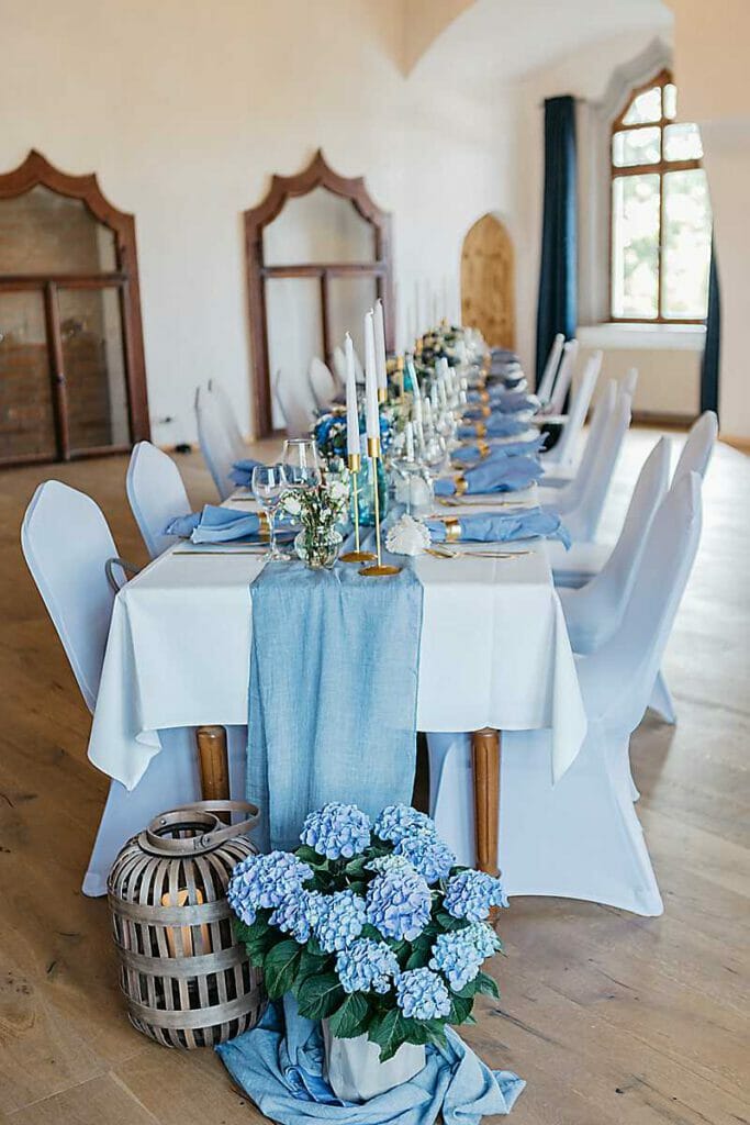 Elegant gedeckter Esstisch für eine Veranstaltung einer Hochzeitsplanerin in Leipzig und Usedom mit weißen Stühlen, blauem Tischläufer, Kerzen und Glaswaren. Blaue Hortensien und eine Weidenlaterne schmücken den lichtdurchfluteten Raum mit Rundbogenfenstern.