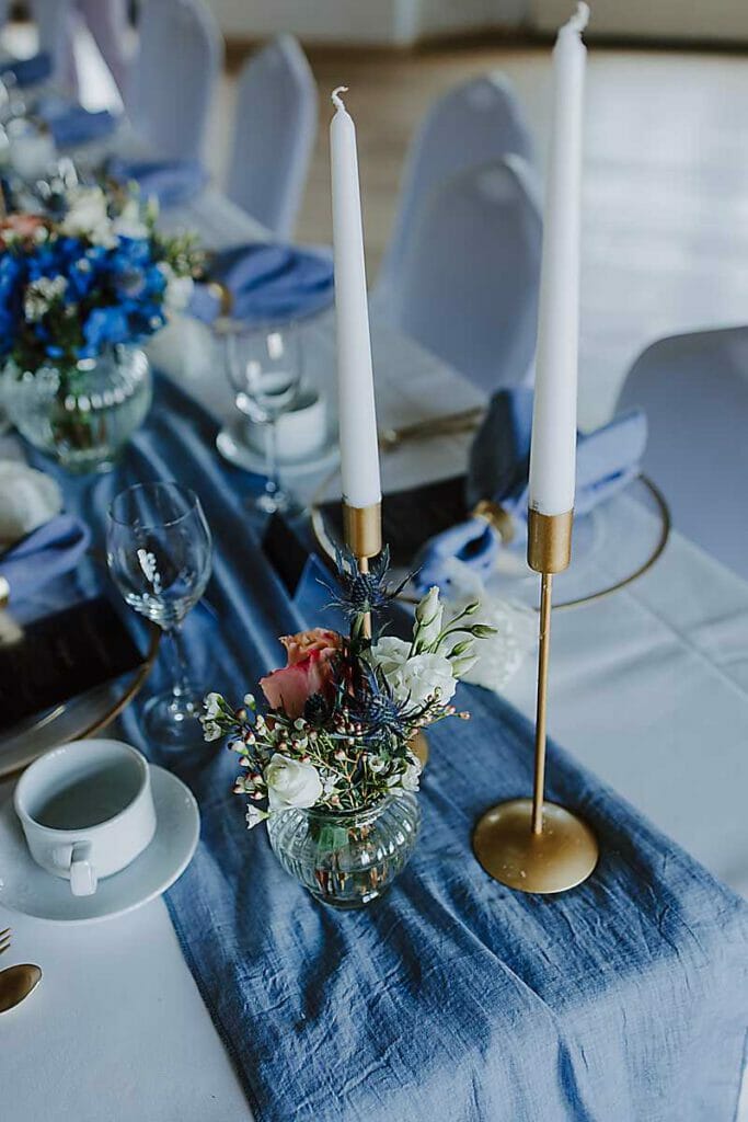 Nahaufnahme eines elegant gedeckten Tisches einer Hochzeitsplanerin in Leipzig und Usedom mit zwei hohen weißen Kerzen in goldenen Haltern, einer kleinen Glasvase mit Blumen, blauem Tischläufer, weißen Tellern, Tassen, Gläsern und ordentlich gefalteten Servietten.