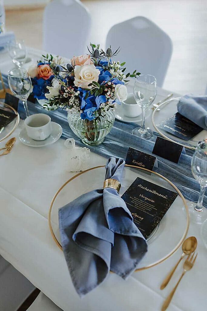 Elegant gedeckter Tisch mit goldumrandeten Tellern, blauen Servietten, goldenem Besteck, Weingläsern, einer Menükarte und einem floralen Mittelstück - perfekt kuratiert von einer Hochzeitsplanerin in Leipzig und Usedom für Ihren besonderen Tag.