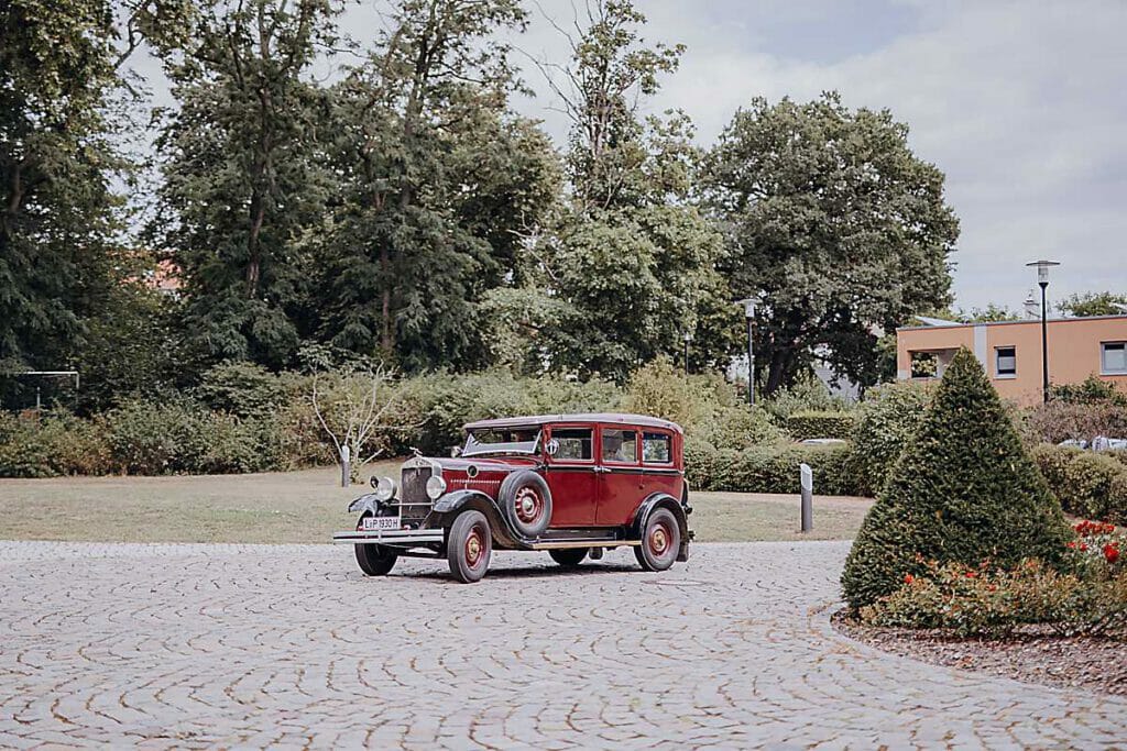 Ein roter Oldtimer mit weißwandigen Reifen steht auf einer kopfsteingepflasterten Auffahrt, umgeben von Grün und Bäumen - perfekt für eine Hochzeitsplanerin in Leipzig und Usedom, die eine elegante Hochzeit in der Nähe eines modernen Gebäudes plant.