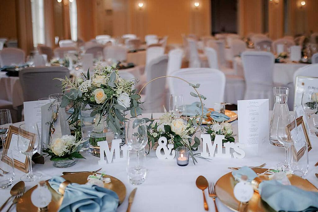 Eine Hochzeitstafel, dekoriert mit weißem und goldenem Geschirr, eleganten Blumenarrangements, einem Mr & Mrs Schild, Speisekarten und Bilderrahmen - gestaltet von einer Hochzeitsplanerin in Leipzig und Usedom. Der Hintergrund leuchtet mit sanft beleuchteten Tischen und weißen Stühlen.