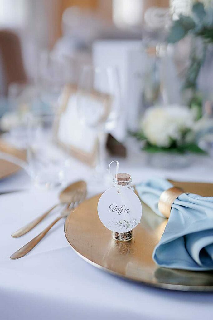 Nahaufnahme eines elegant gedeckten Tisches, den eine Hochzeitsplanerin in Leipzig und Usedom arrangiert hat, mit einem goldenen Platzteller, einer blauen Serviette, einem silbernen Besteck und einer Tischkarte aus Glasflaschen mit der Aufschrift Stefan. Der Hintergrund ist sanft verschwommen.