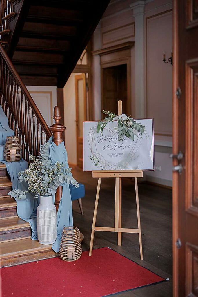Ein Hochzeitsbegrüßungsschild auf einer Staffelei, arrangiert von einer Hochzeitsplanerin in Leipzig und Usedom, ist mit Grünpflanzen neben einer mit blauem Stoff drapierten Treppe, Weidenlaternen und Blumenvasen auf einem roten Teppich in einem eleganten Innenbereich geschmückt.