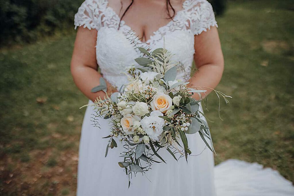 Eine Braut in einem weißen Spitzenhochzeitskleid hält einen Strauß weißer und hellgelber Blumen mit grünem Laub und steht im Freien auf einer Wiese - die perfekte Inspiration für jede Hochzeitsplanerin in Leipzig und Usedom.