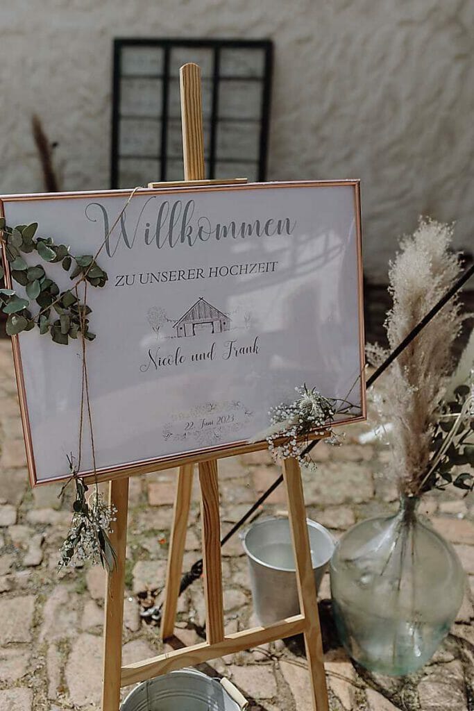 Ein Hochzeitsbegrüßungsschild auf einer Staffelei lautet: "Willkommen zu unserer Hochzeit, Nicole und Frank, 23. Juni 2023". Es wurde von einer Hochzeitsplanerin in Leipzig und Usedom entworfen und zeigt Grünpflanzen, rustikales Dekor und frische Blumen auf Kopfsteinpflaster.