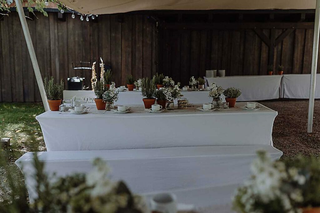 Lange weiße Tische mit eleganten Tischdecken sind für eine Veranstaltung im Freien gedeckt, mit zarten Blumenarrangements und Topfpflanzen. Die Umgebung, die von einem Vordach neben einem Holzgebäude beschattet wird, spiegelt den Stil einer erfahrenen Hochzeitsplanerin in Leipzig und Usedom wider.