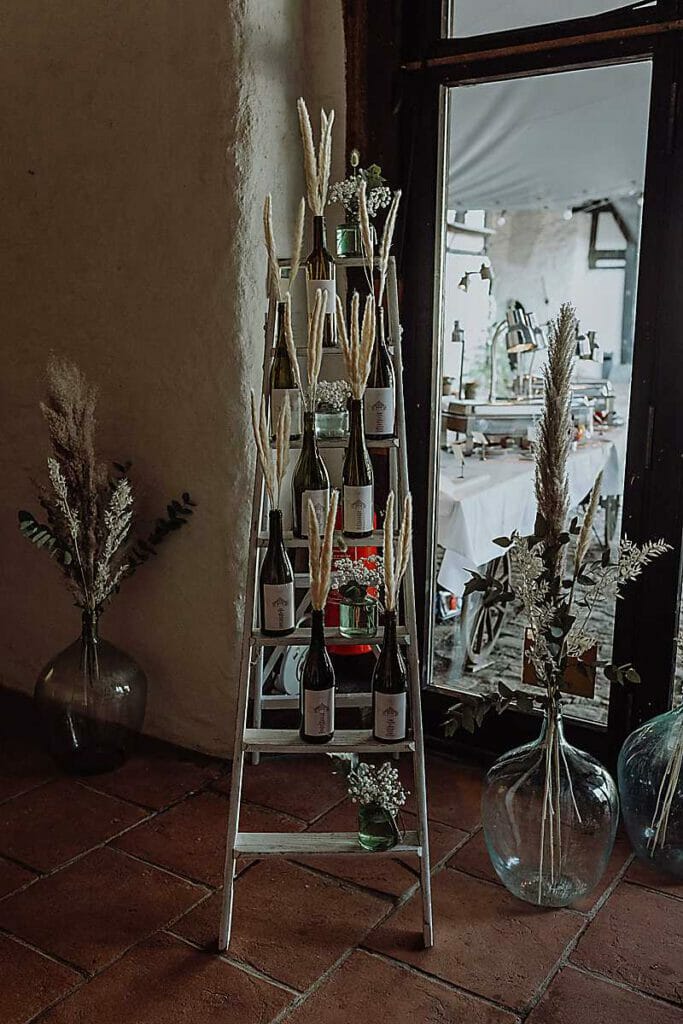 Auf einer dekorativen Holzleiter, die von einer Hochzeitsplanerin in Leipzig und Usedom gestaltet wurde, stehen mit getrocknetem Pampasgras gefüllte Weinflaschen neben großen Glasvasen und rustikalen Blumenarrangements an einem sonnendurchfluteten Fenster.