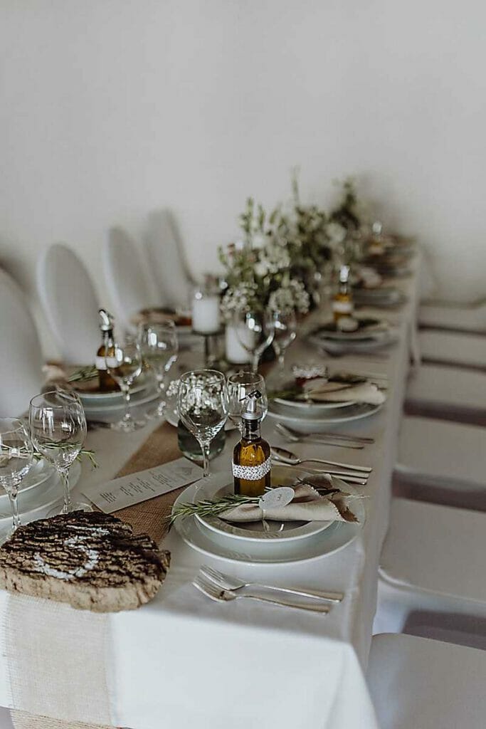 Ein langer Esstisch, gedeckt für ein formelles Essen mit weißen Stühlen, Tellern, Weingläsern, rustikalem Dekor, weißer Bettwäsche und einem Mittelstück aus Grünzeug und Blumen - eine perfekte Inspiration von einer Hochzeitsplanerin in Leipzig und Usedom.