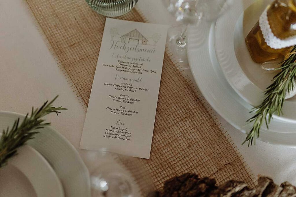 Ein Hochzeitsmenü auf einem rustikalen Tisch mit weißen Tellern, einem Rosmarinzweig, einer Flasche mit Spitzenband und einem Läufer aus Sackleinen - gestaltet von einer Hochzeitsplanerin in Leipzig und Usedom. Die Speise- und Getränkekarte ist in eleganter Schrift verfasst.