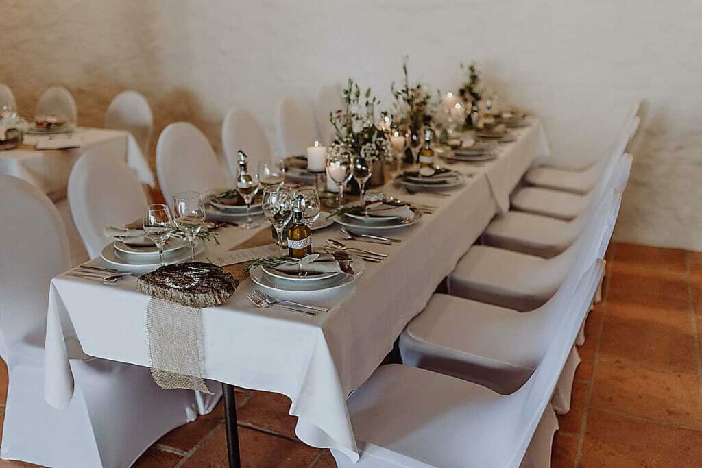 Ein langer Esstisch, der von einer Hochzeitsplanerin in Leipzig und Usedom für eine feierliche Veranstaltung gedeckt wurde, mit weißen Tischtüchern, eleganten Stühlen, Tellern, Gläsern, Kerzen, Topfpflanzen und Olivenölflaschen, fein säuberlich angeordnet.