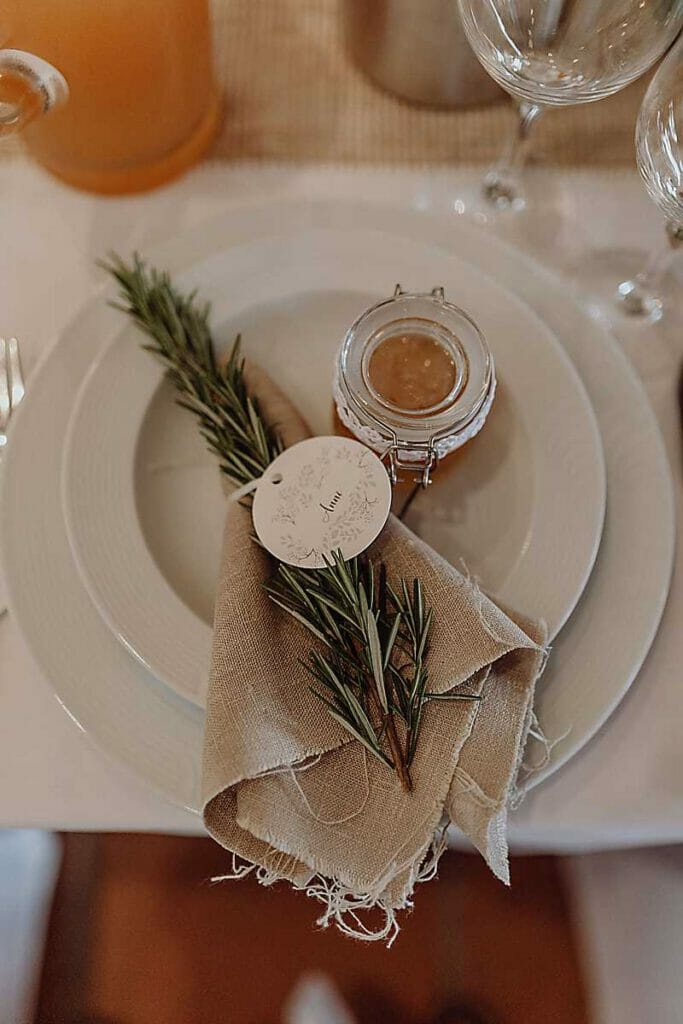 Auf dem gedeckten Tisch einer Hochzeitsplanerin in Leipzig und Usedom stehen ein weißer Teller, eine beigefarbene Serviette mit Rosmarin und ein Namensschild sowie ein Glas mit goldbraunem Brotaufstrich. Weingläser und ein Krug vervollständigen die elegante Szene.