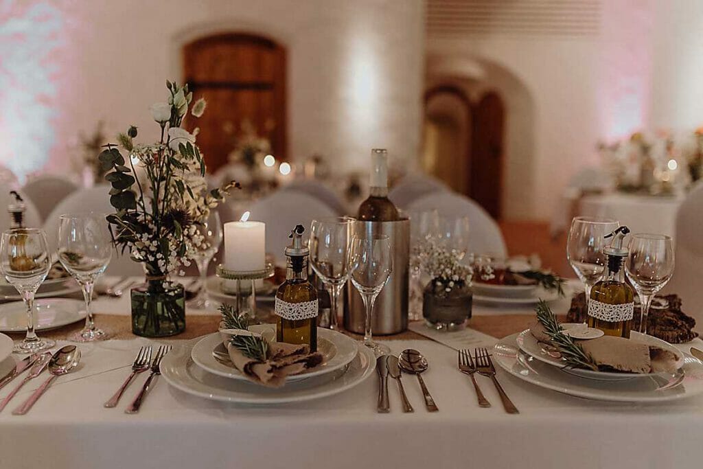 Ein wunderschön dekorierter Hochzeitstisch mit Tellern, Besteck, Weingläsern, Kerzen, kleinen Blumenarrangements und Olivenölflaschen - gestylt von einer Hochzeitsplanerin in Leipzig und Usedom - mit sanfter Beleuchtung und einem unscharfen, eleganten Veranstaltungshintergrund.