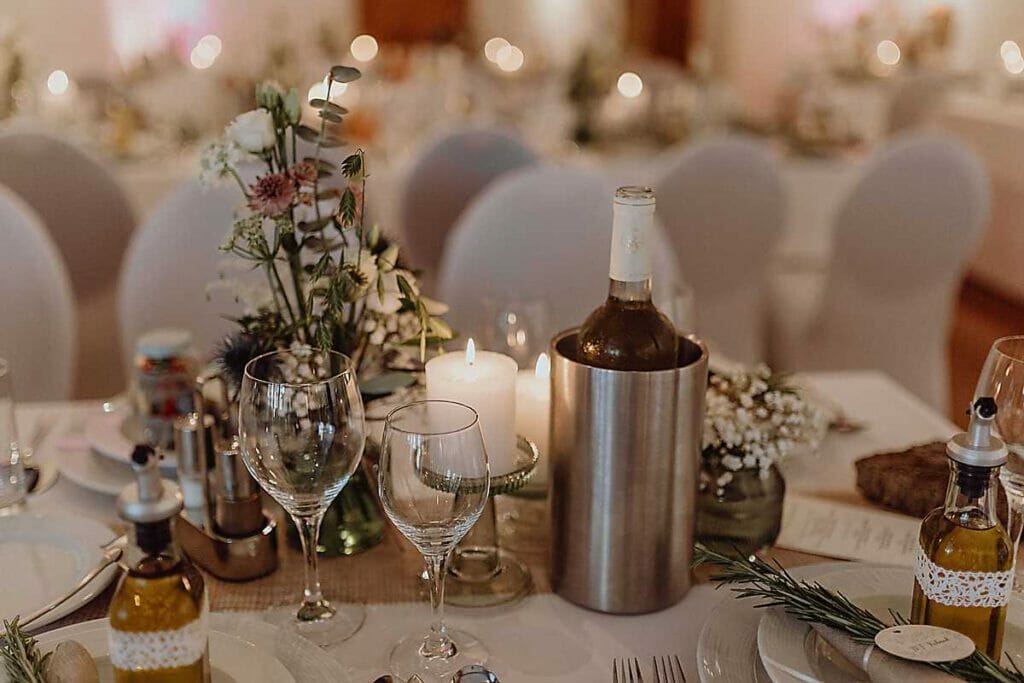 Ein geschmückter Hochzeitstisch mit Weingläsern, Kerzen, floralen Tafelaufsätzen, einer Weinflasche im Kühler, Olivenölflaschen und eleganten weißen Stühlen - fachmännisch gestylt von Ihrer Hochzeitsplanerin in Leipzig und Usedom.