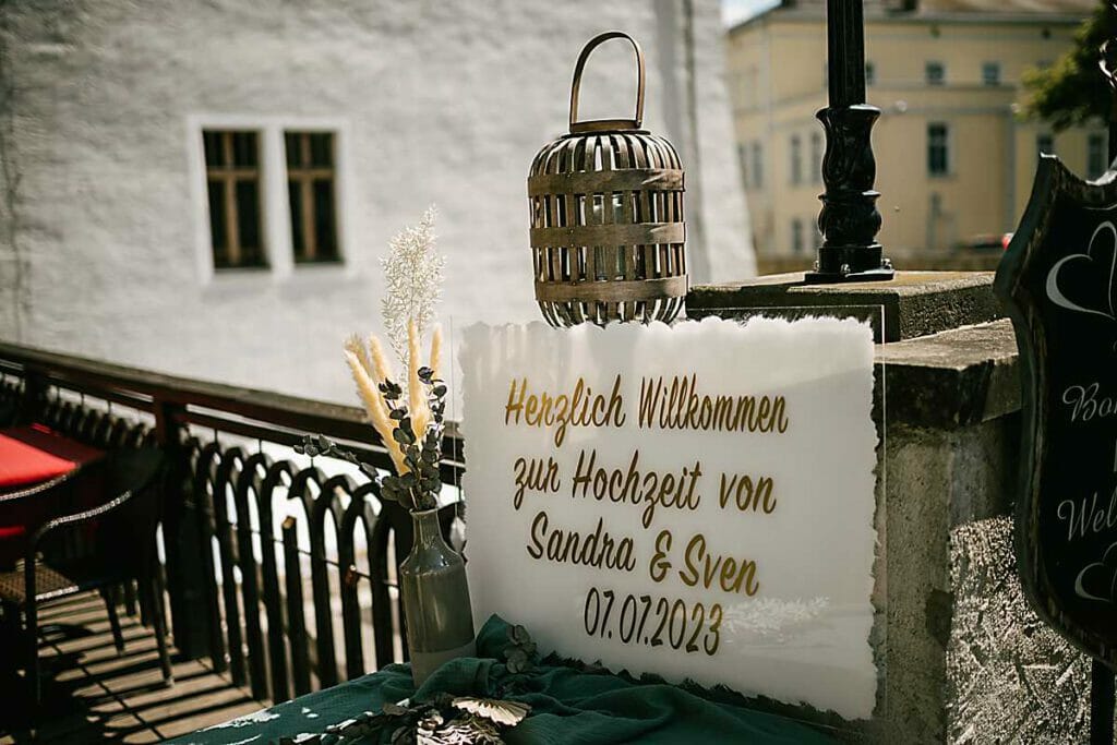 Ein Hochzeits-Willkommensschild mit der Aufschrift "Herzlich Willkommen zur Hochzeit von Sandra & Sven 07.07.2023" steht im Freien mit Dekorationsartikeln auf einem Tisch und einem weißen Gebäude im Hintergrund - perfekt gestaltet von einer Hochzeitsplanerin in Leipzig und Usedom.