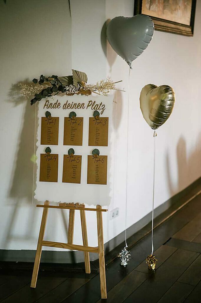 Ein Hochzeitssitzplan auf einer Staffelei, dekoriert mit Trockenblumen und zwei herzförmigen Luftballons (einer silber, einer gold) in der Nähe. Perfekt arrangiert von einer Hochzeitsplanerin in Leipzig und Usedom, listet die Karte sechs Tische unter "Finde deinen Platz" auf.
