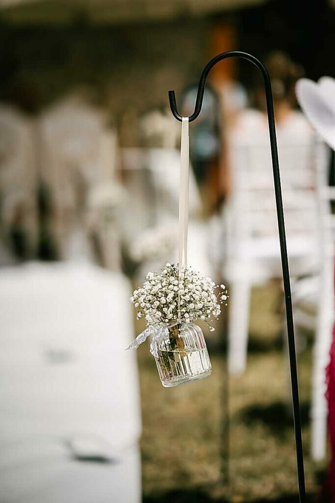 Ein kleines Glasgefäß, gefüllt mit zarten weißen Schleierkrautblüten, hängt an einem schwarzen Metallhaken, wahrscheinlich als Hochzeitsdekoration im Freien, gestylt von einer Hochzeitsplanerin in Leipzig und Usedom. Im Hintergrund sind weiße Stühle und unscharfe Gäste zu sehen.