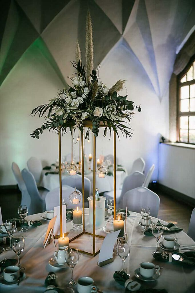 Elegante Hochzeitstischdekoration mit einem hohen goldenen Rahmen, der üppige Blumen und hängende Glaskugeln mit Kerzen hält, entworfen von einer Hochzeitsplanerin in Leipzig und Usedom, umgeben von weiteren Kerzen und schickem Geschirr in einem sanft beleuchteten, modernen Raum.