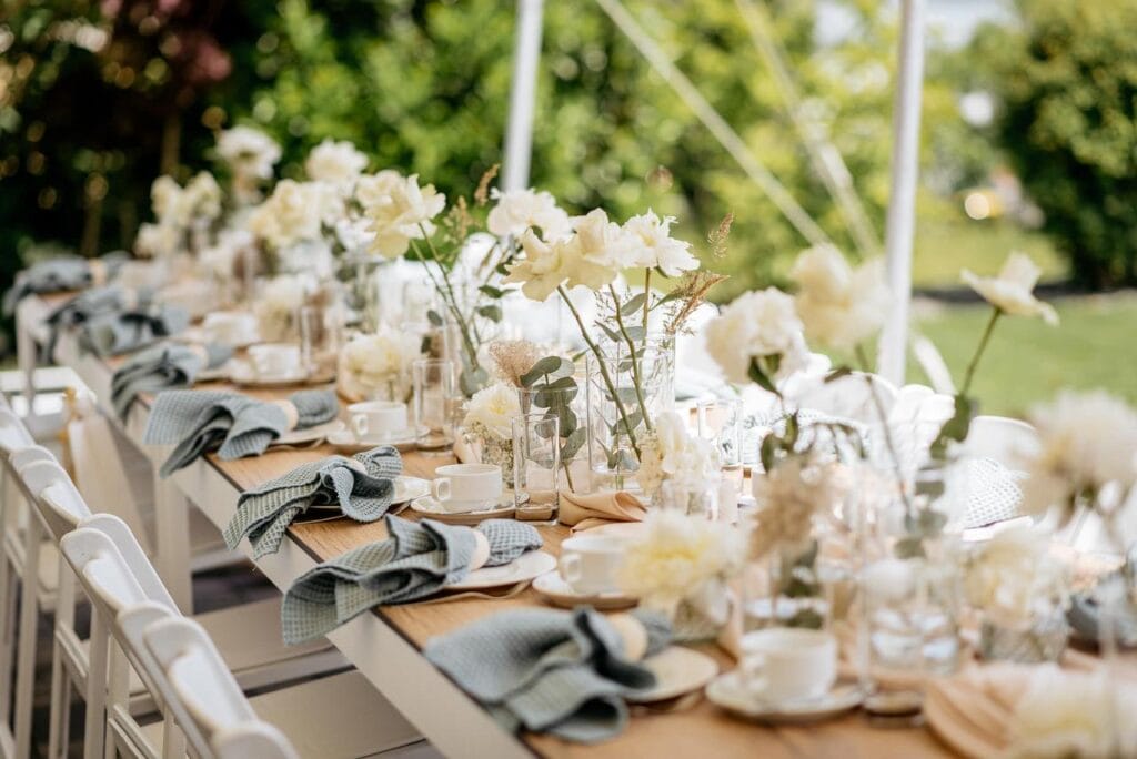 Ein langer Tisch im Freien ist elegant mit weißem Geschirr, hellblauen Servietten, Gläsern und weißen Blumen in der Mitte gedeckt - eine exquisite Szene, die von einer Hochzeitsplanerin in Leipzig und Usedom arrangiert wurde, perfekt für eine formelle Zusammenkunft inmitten von natürlichem Licht und Grün.