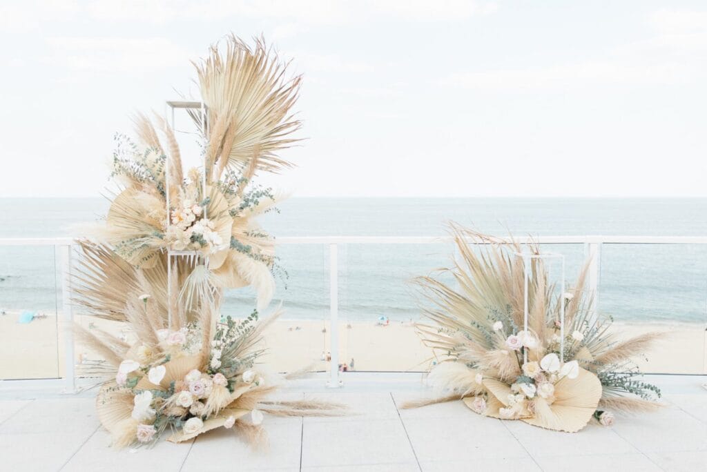 Zwei große Trockenblumenarrangements mit Pampasgras, Palmenblättern und hellen Blumen schmücken eine Terrasse mit Blick auf einen Sandstrand - eine verträumte Szenerie, inspiriert von einer Hochzeitsplanerin in Leipzig und Usedom unter strahlendem Himmel.