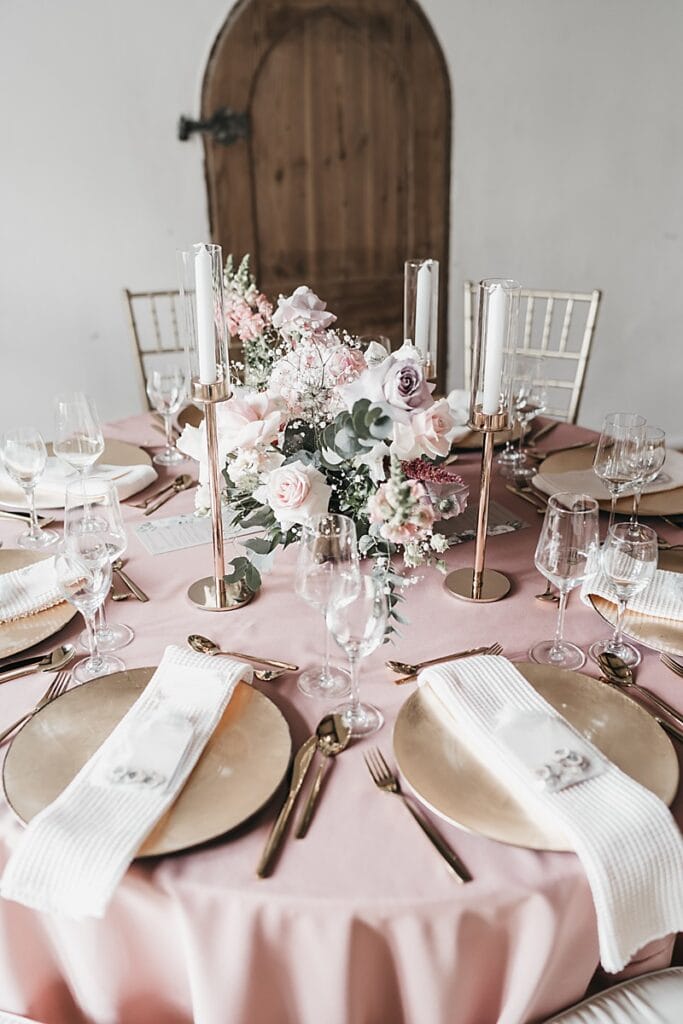 Ein runder Tisch mit rosafarbener Tischdecke ist für vier Personen gedeckt, mit goldenen Tellern, goldenem Besteck, klaren Gläsern, elfenbeinfarbenen Servietten und einem Blumenschmuck aus rosafarbenen und weißen Blumen, mit hohen Kerzenleuchtern und einer Holztür im Hintergrund.