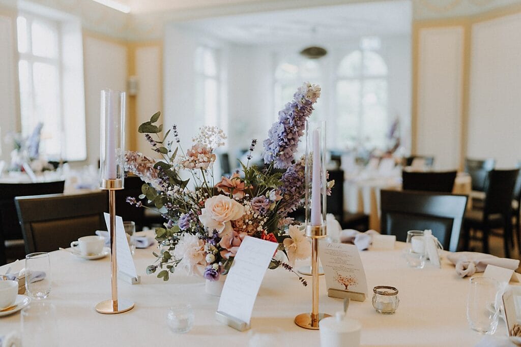 Eleganter Hochzeitstisch mit einem Mittelstück aus pastellfarbenen Blumen, hohen Glaskerzenhaltern mit goldenen Sockeln, Menüs, Tassen und Gläsern auf einem weißen Tischtuch in einem hellen, luftigen Raum.