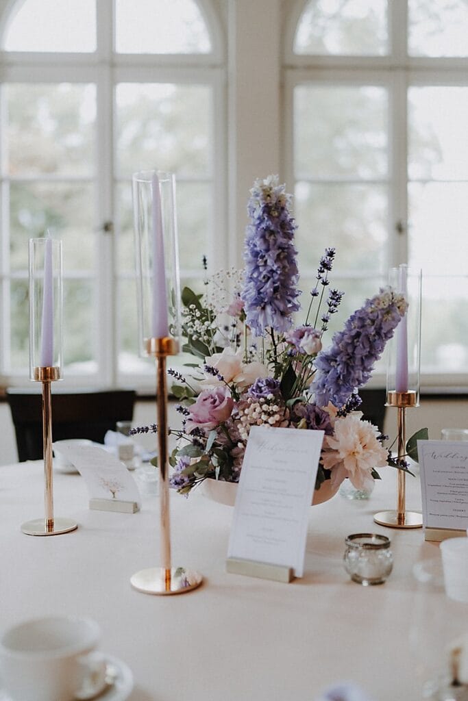 Ein Tafelaufsatz mit hohen Lavendelkerzen in goldenen Haltern, ein Blumenarrangement aus violetten, weißen und rosafarbenen Blumen und elegante Menüs auf einer hellen Tischdecke vor großen Bogenfenstern.