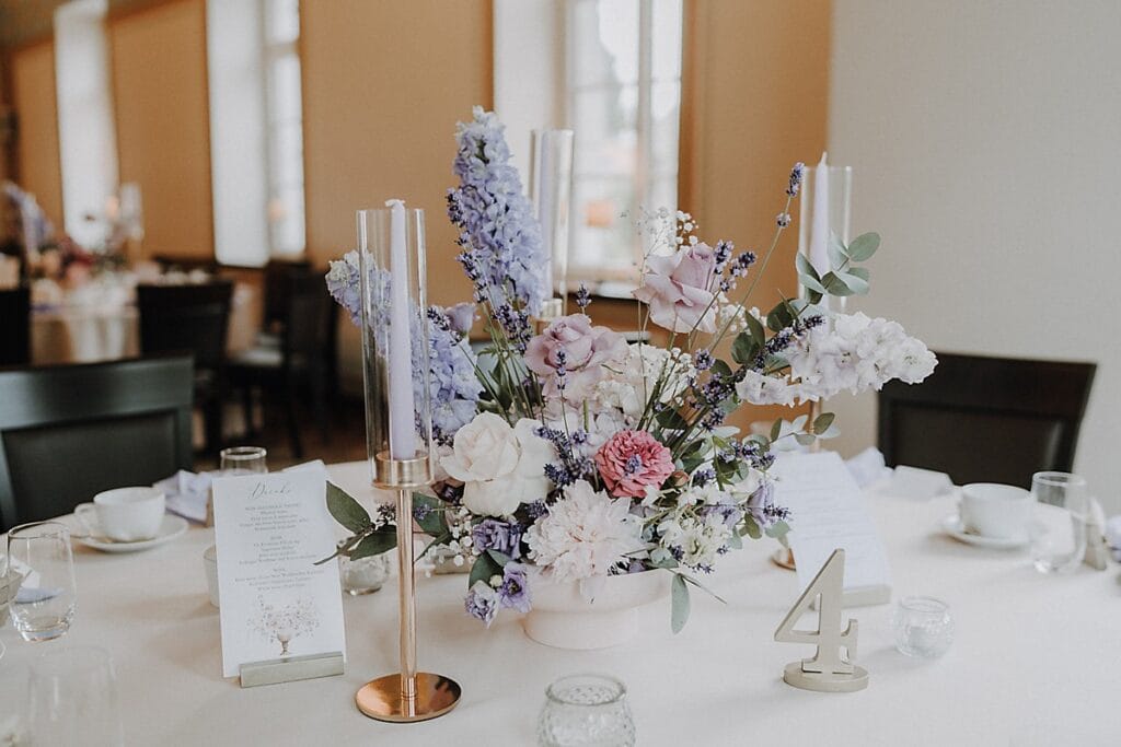 Ein für eine Veranstaltung gedeckter runder Tisch mit einem Mittelstück aus pastellfarbenen Blumen, hohen Kerzen in Glashaltern, einer Menükarte und einer metallischen Tischnummer 4. Im Hintergrund sind Fenster und andere, ähnlich gedeckte Tische zu sehen.