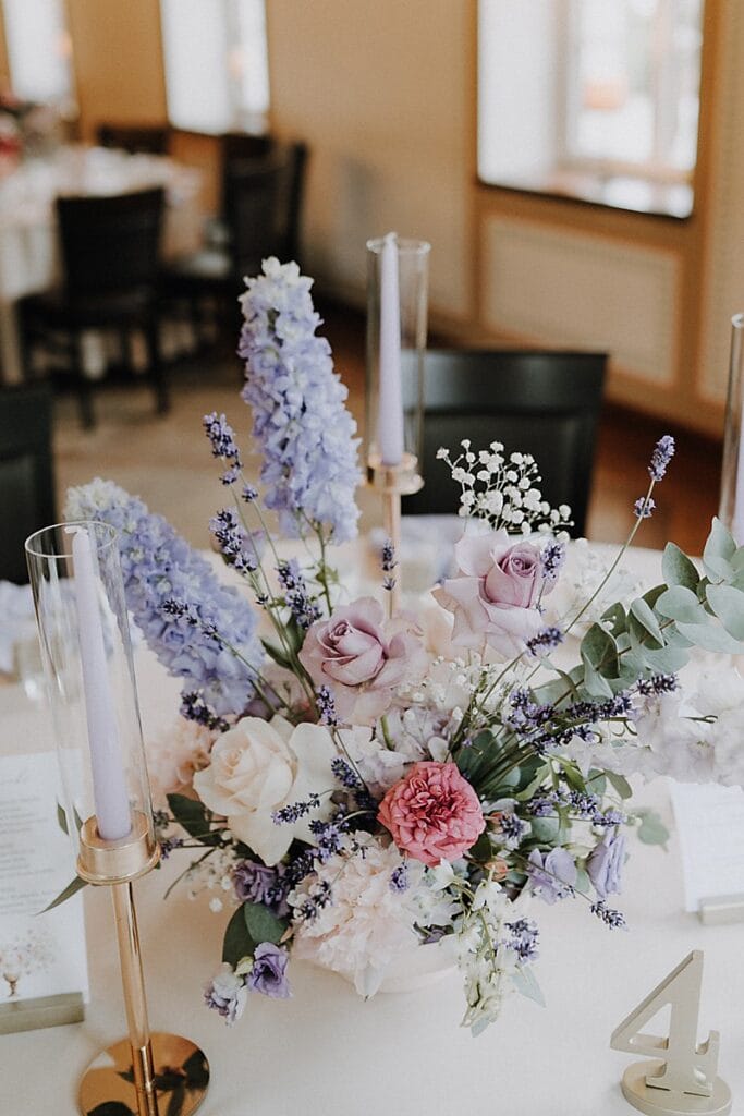 Elegantes florales Mittelstück mit lavendelfarbenen, rosafarbenen und weißen Blumen, darunter Rosen und Rittersporn, umgeben von hohen, hellvioletten Kerzen auf goldenen Haltern, auf einem runden Tisch mit schwarzen Stühlen in einem sanft beleuchteten Raum.