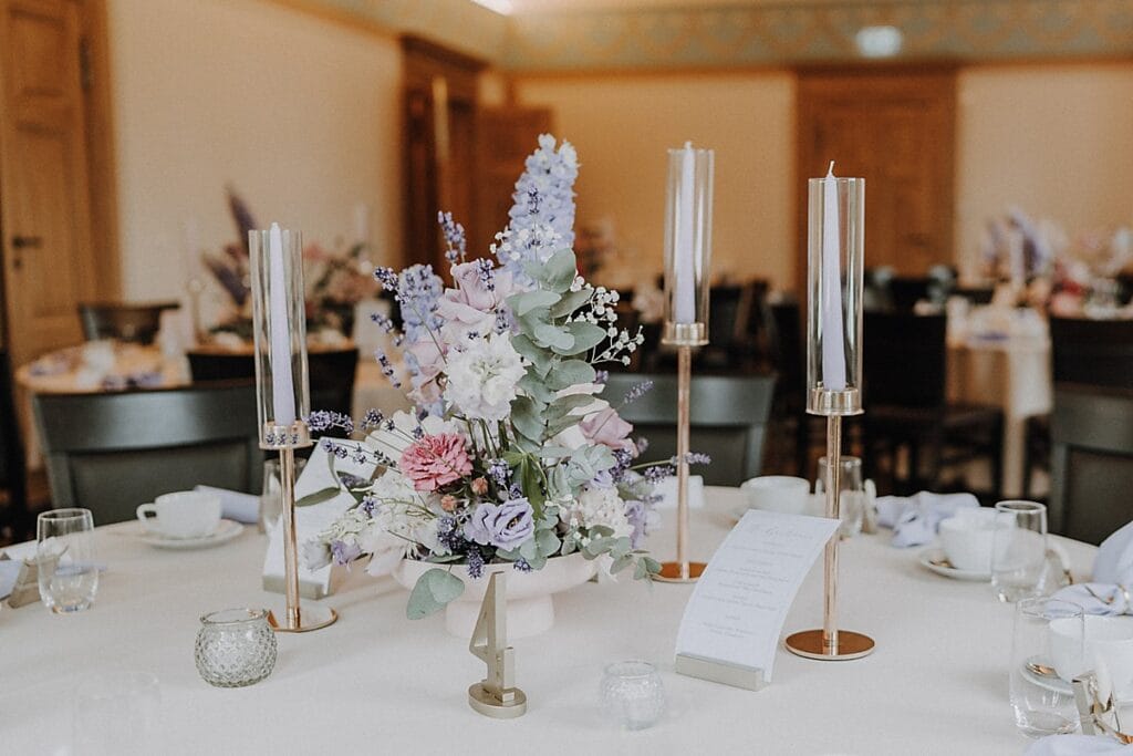 Elegant gedeckter Tisch mit einem Mittelstück aus pastellfarbenen Blumen und drei hohen Kerzenhaltern aus Glas. Um das Arrangement herum stehen Gläser, Tassen, eine Speisekarte und kleine Dekorationen auf einem weißen Tischtuch.