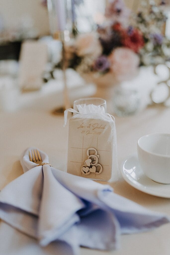 Ein Hochzeitstisch mit einer lavendelfarbenen Serviette, einer goldenen Gabel, einer weißen Teetasse und einer Geschenktüte mit einem Herzmotiv und den Namen Sara & Lukas mit Datum, mit verschwommenen Blumen im Hintergrund.