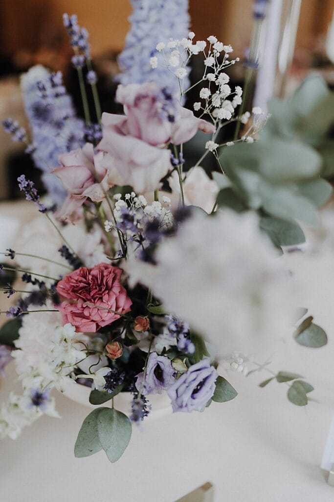 Eine Nahaufnahme eines Blumenarrangements mit rosa, lavendelfarbenen und weißen Blumen mit Grünzeug und zarten Zweigen von Babyatem in einer weißen Vase auf einer hellen Fläche.