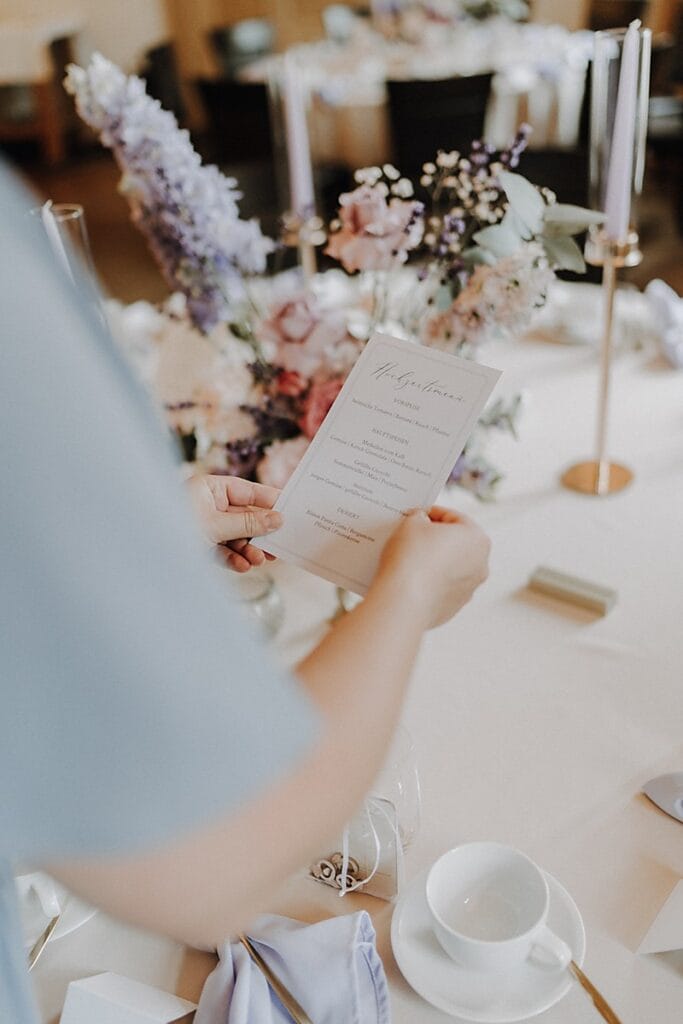 Eine Person in hellblauem Outfit hält ein Hochzeitsmenü an einem elegant gedeckten Tisch mit Blumenschmuck, Kerzen, weißem Geschirr und Dekorationen in zarten Pastelltönen.