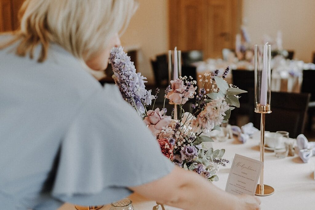 Eine Frau arrangiert einen Blumenschmuck mit rosafarbenen und violetten Blumen auf einem für eine Veranstaltung gedeckten Tisch, auf dem Kerzen und eine gedruckte Karte in der eleganten Umgebung zu sehen sind.