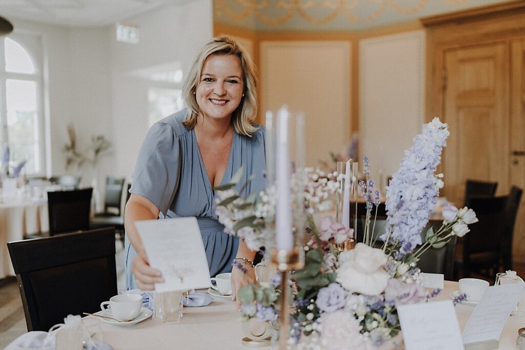 Eine lächelnde Frau in einem hellblauen Kleid steht an einem elegant gedeckten Tisch mit fliederfarbenen und weißen Blumen, lila Kerzen und Speisekarten in einem hellen, dekorierten Raum. Sie hält eine Menükarte in der Hand und blickt in die Kamera.