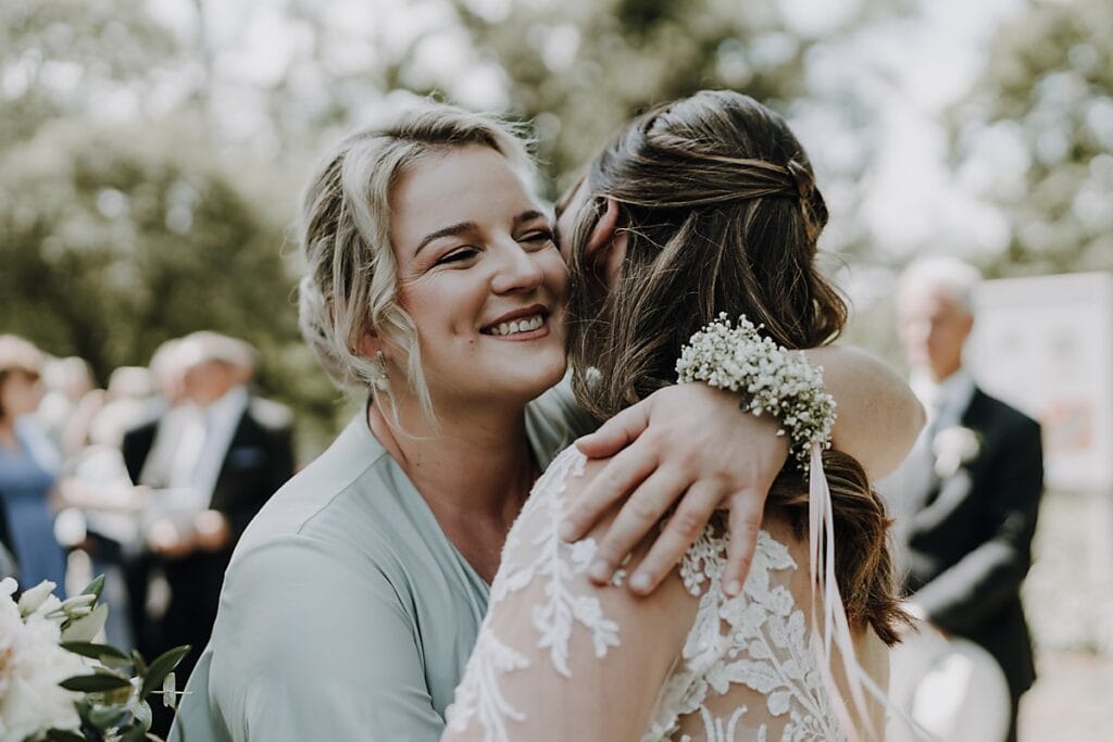 Zwei Frauen umarmen sich im Freien bei einer Hochzeit. Die eine Frau trägt lächelnd ein hellgrünes Kleid, die andere ein weißes Spitzenkleid und eine Blumenkorsage. Menschen und Grünpflanzen sind im Hintergrund verschwommen.