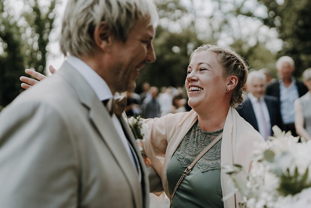 Eine lächelnde Frau begrüßt einen Mann in einem hellen Anzug im Freien bei einer Veranstaltung, möglicherweise einer Feier oder Hochzeit, mit unscharfen Menschen und Grün im Hintergrund.