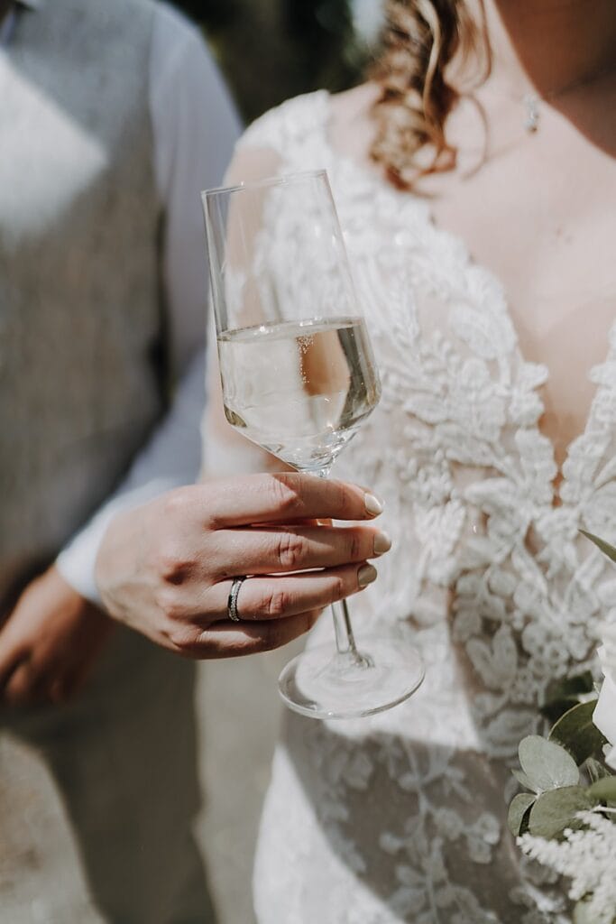 Eine Braut in einem weißen Spitzenkleid hält ein Glas Champagner und zeigt ihren Verlobungsring. Das Foto ist eine Nahaufnahme, auf der ein Teil einer anderen Person neben ihr zu sehen ist.