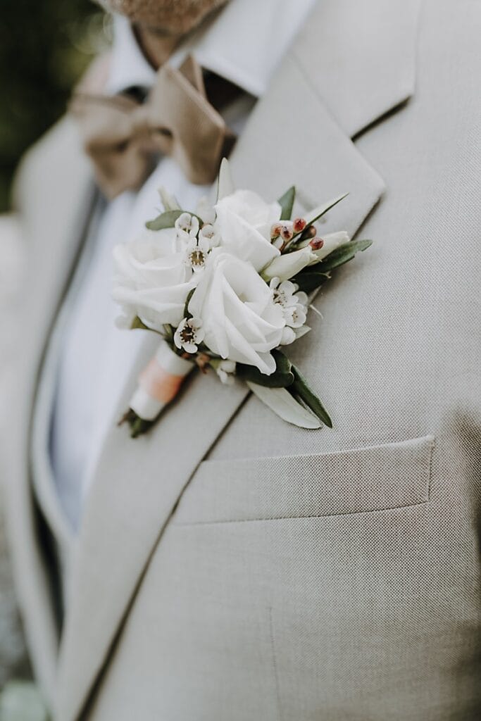 Nahaufnahme einer Person, die einen hellbeigen Anzug mit einer braunen Fliege trägt, an deren Revers eine weiße Rosenboutonniere befestigt ist. Der Schwerpunkt liegt auf den Details des Anzugs und dem Blumenarrangement.
