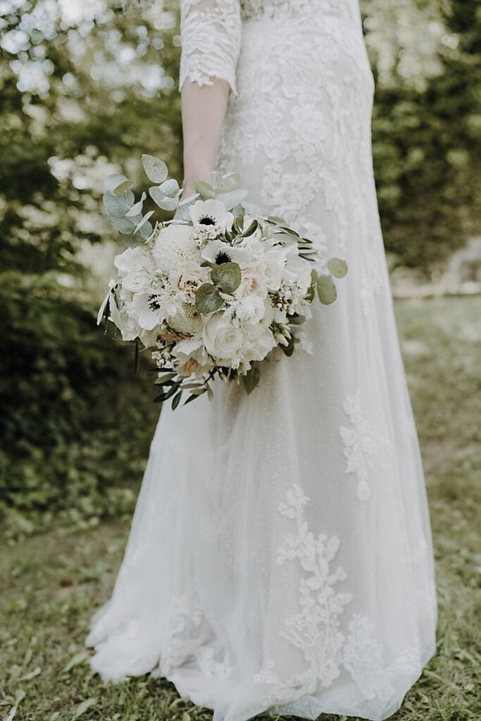 Eine Braut im Spitzenhochzeitskleid steht im Freien und hält einen Strauß aus weißen und cremefarbenen Blumen mit Grünzeug. Das Bild konzentriert sich auf die untere Hälfte ihres Kleides und den Blumenstrauß, mit einem unscharfen grünen Hintergrund.