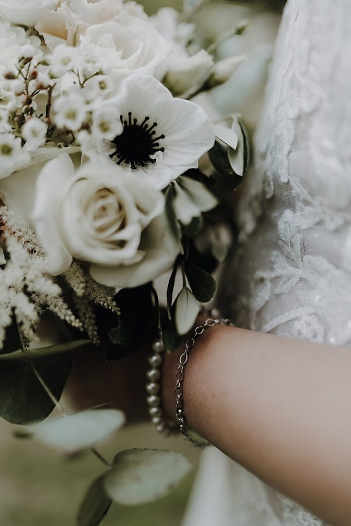 Nahaufnahme der Hand einer Braut, die einen Strauß weißer Blumen, darunter Rosen und Anemonen, mit Grünzeug hält. Die Braut trägt Perlen- und Kettenarmbänder, und ihr Spitzenhochzeitskleid ist teilweise sichtbar.