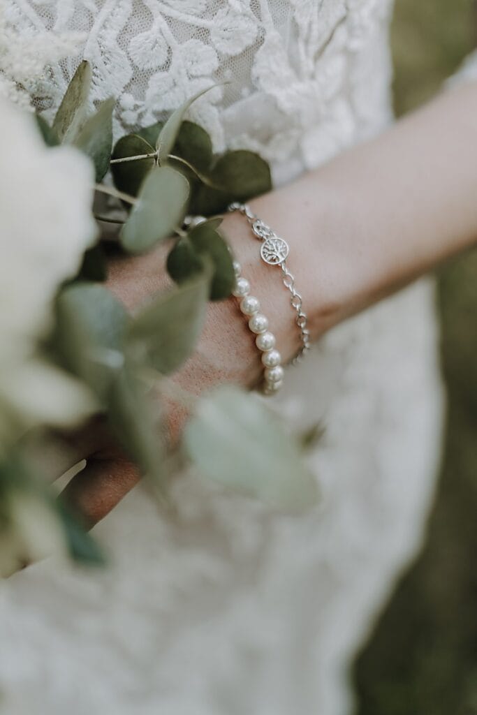 Nahaufnahme einer Braut, die einen grünen Strauß in der Hand hält und ein zartes Perlenarmband und ein silbernes Armband mit einem Anhänger trägt. Im Hintergrund ist ihr Hochzeitskleid aus Spitze zu sehen.