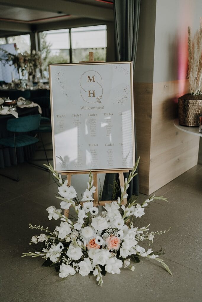 Ein Sitzplan für eine Hochzeit auf einer Staffelei, dekoriert mit einem Blumenarrangement aus weißen und rosafarbenen Blumen an der Basis. Die Karte ist mit eleganten Buchstaben und einem herzlichen Willkommensgruß versehen und befindet sich in einem modernen Innenbereich.