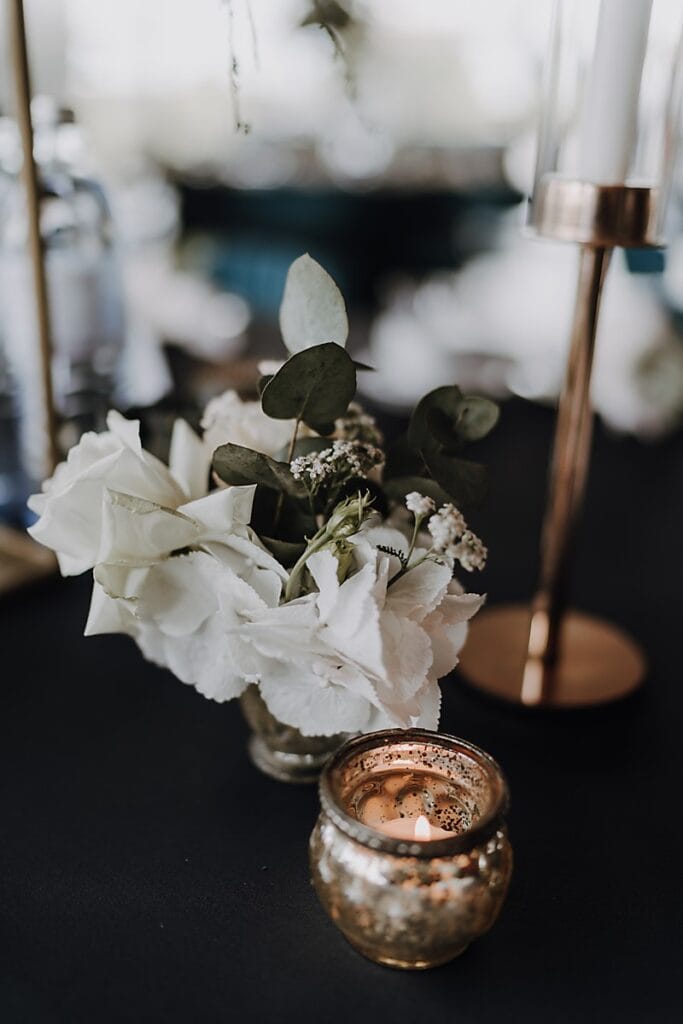 Ein kleiner Strauß weißer Blumen und grüner Blätter steht in einer Glasvase neben einem beleuchteten goldenen Kerzenständer auf einer dunklen Tischplatte, mit einem verschwommenen Kerzenständer und Flaschen im Hintergrund.