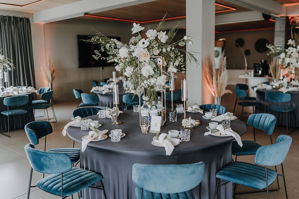 Ein runder Tisch mit dunklem Tischtuch ist elegant für eine Veranstaltung gedeckt, auf dem ein hoher floraler Tafelaufsatz mit weißen Blumen und Grünzeug steht. Blaue Samtstühle umgeben den Tisch in einem modernen, stilvollen Raum.