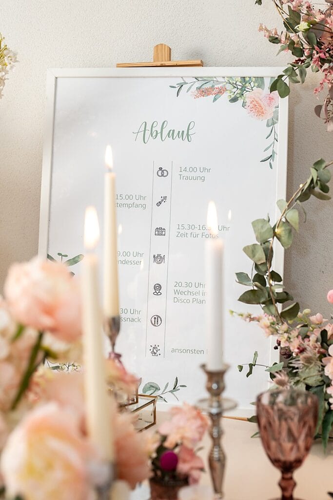 Ein gerahmter Hochzeitsterminplan in deutscher Sprache wird auf einer Staffelei ausgestellt, umgeben von rosa Blumen und brennenden Kerzen, mit eleganten dekorativen Elementen im Vordergrund. Der Zeitplan listet Ereignisse und Zeiten auf, mit floralen Akzenten auf dem Rahmen.