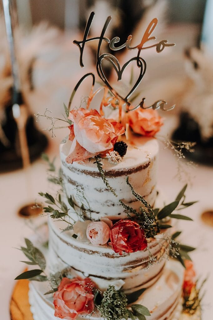 Eine dreistöckige, halbnackte Hochzeitstorte, verziert mit rosa und roten Blumen und Grünzeug, gekrönt von einem Metallschild mit der Aufschrift Herb ♥ Maria.