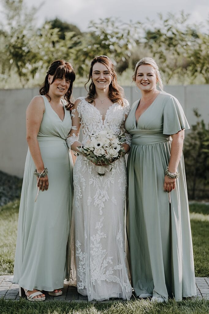 Drei Frauen stehen im Freien zusammen. Die Frau in der Mitte trägt ein weißes Hochzeitskleid und hält einen Blumenstrauß. Die beiden Frauen neben ihr tragen passende hellgrüne Kleider. Alle lächeln und stehen auf einer Wiese mit grünem Bewuchs im Hintergrund.