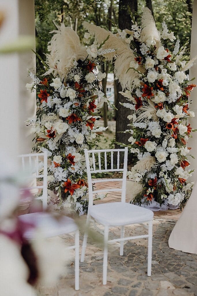 Zwei weiße Stühle stehen vor einem blumengeschmückten Hochzeitsbogen mit weißen Rosen, orangefarbenen Lilien, Grünzeug und Pampasgras, der im Freien auf einem Steinweg in der Nähe von Bäumen aufgestellt ist.