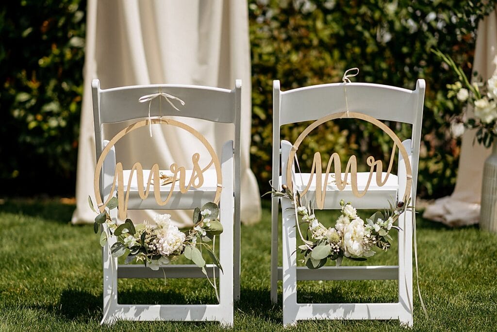 Zwei weiße Stühle im Gras, dekoriert mit runden "Mrs"- und "Mr"-Schildern, Grünzeug und weißen Blumen. Im Hintergrund sind ein cremefarbener Vorhang und Grünzeug zu sehen, was auf eine Hochzeit hindeutet.